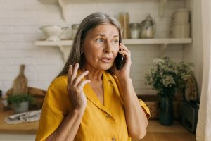 Elderly Caucasian grandmother talking on phone standing at kitchen having anxious and worried face expression